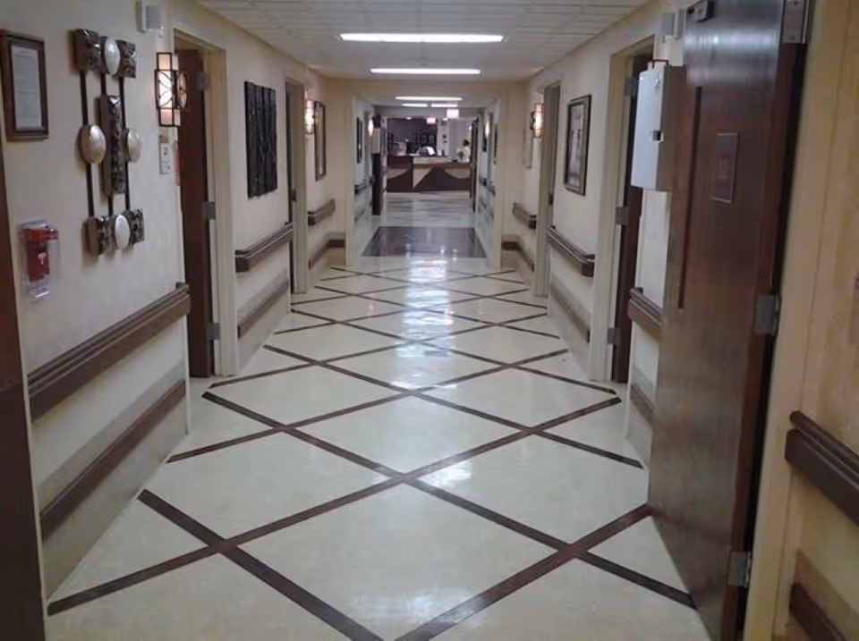 A long, well-lit hallway in a healthcare or rehabilitation facility with patterned flooring, handrails on both sides, wall decorations, and several closed doors leading to rooms. At the end of the hallway, a reception or nurse station is visible.