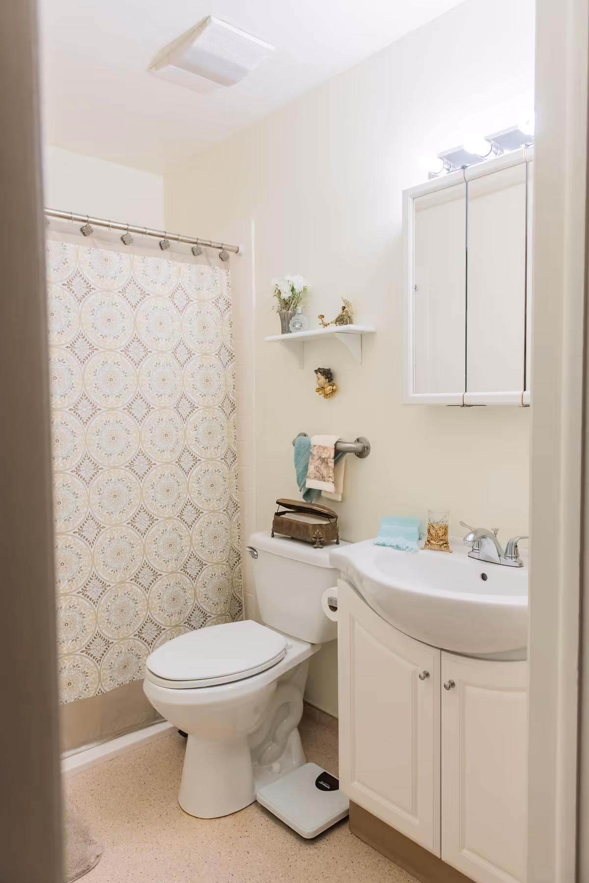 A small bathroom with a white toilet and a white sink cabinet. There is a patterned shower curtain, a small white shelf with decorative items above the toilet, and a mirror with lights above the sink. A scale is placed on the floor in front of the toilet.
