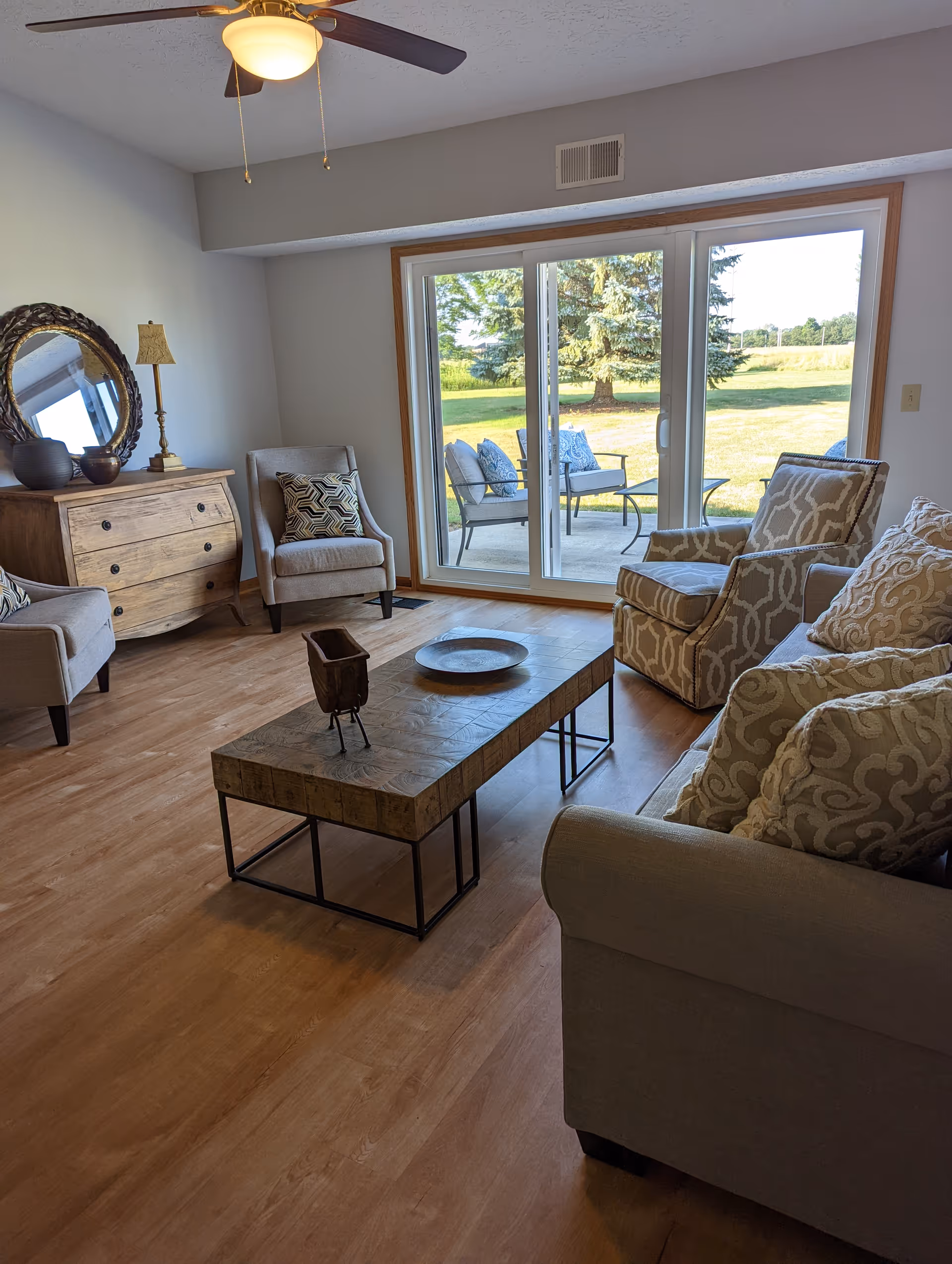 A cozy living room with a beige sofa and patterned armchair, a wooden coffee table with decorative items, a wooden dresser with a round mirror and lamp, and sliding glass doors leading to an outdoor patio with chairs and a table. The room has wooden flooring and a ceiling fan with a light.