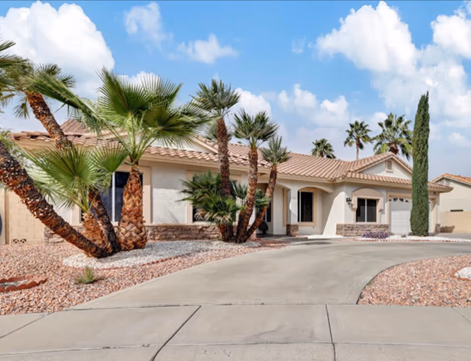 Single-story stucco house with a tile roof, palm trees, and a curved driveway in a desert landscape.