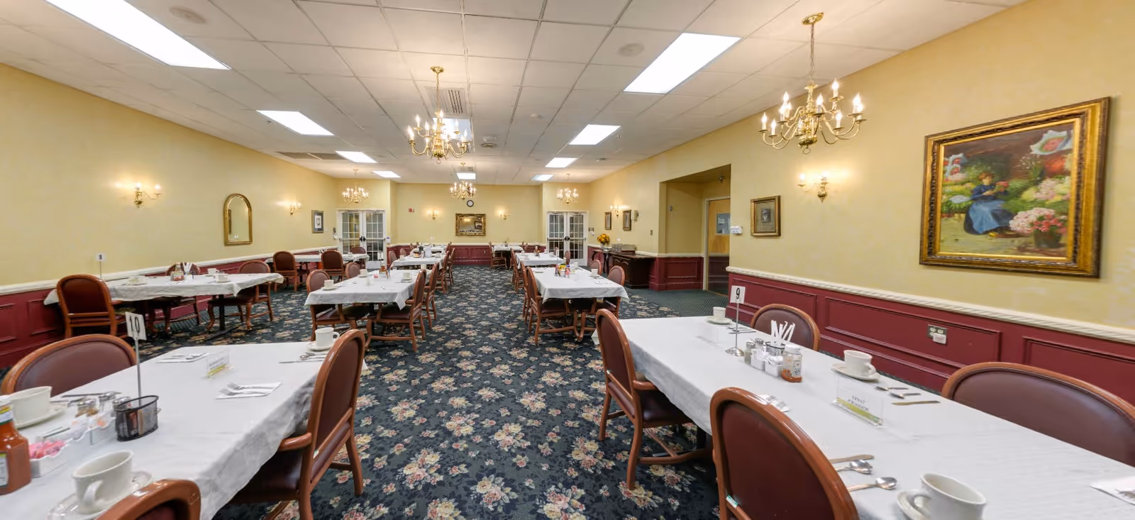 A spacious dining room with multiple tables covered in white tablecloths, set with cups, plates, and condiments. The room features floral carpet, yellow walls with red wainscoting, chandeliers hanging from the ceiling, and framed paintings on the walls.