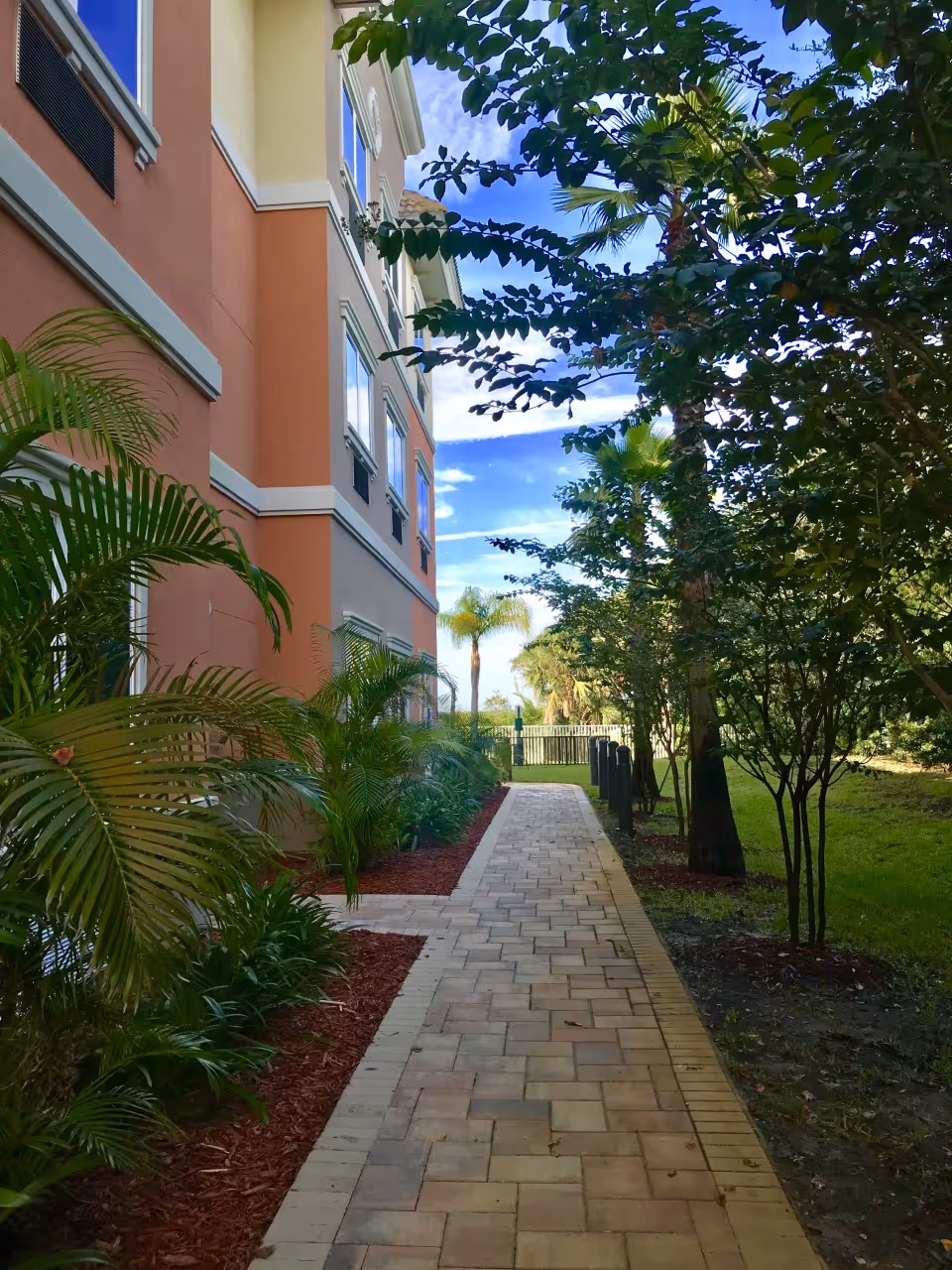 A paved walkway lined with green plants and trees on the right side and a multi-story building with peach and beige walls on the left side under a blue sky with some clouds.