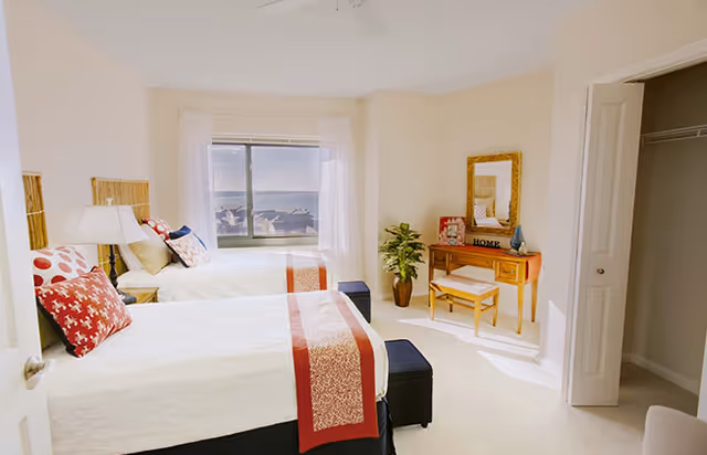 A bright bedroom with two twin beds featuring white bedding and red decorative pillows and runners. There is a window with a view of the water, a wooden vanity table with a mirror, a small bench, a potted plant, and an open door leading to another room.