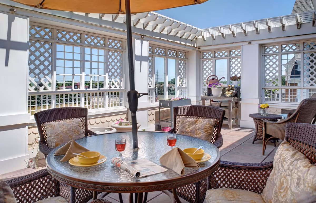 Sunlit outdoor patio with a round glass-top table, wicker chairs, and lattice walls under a pergola.