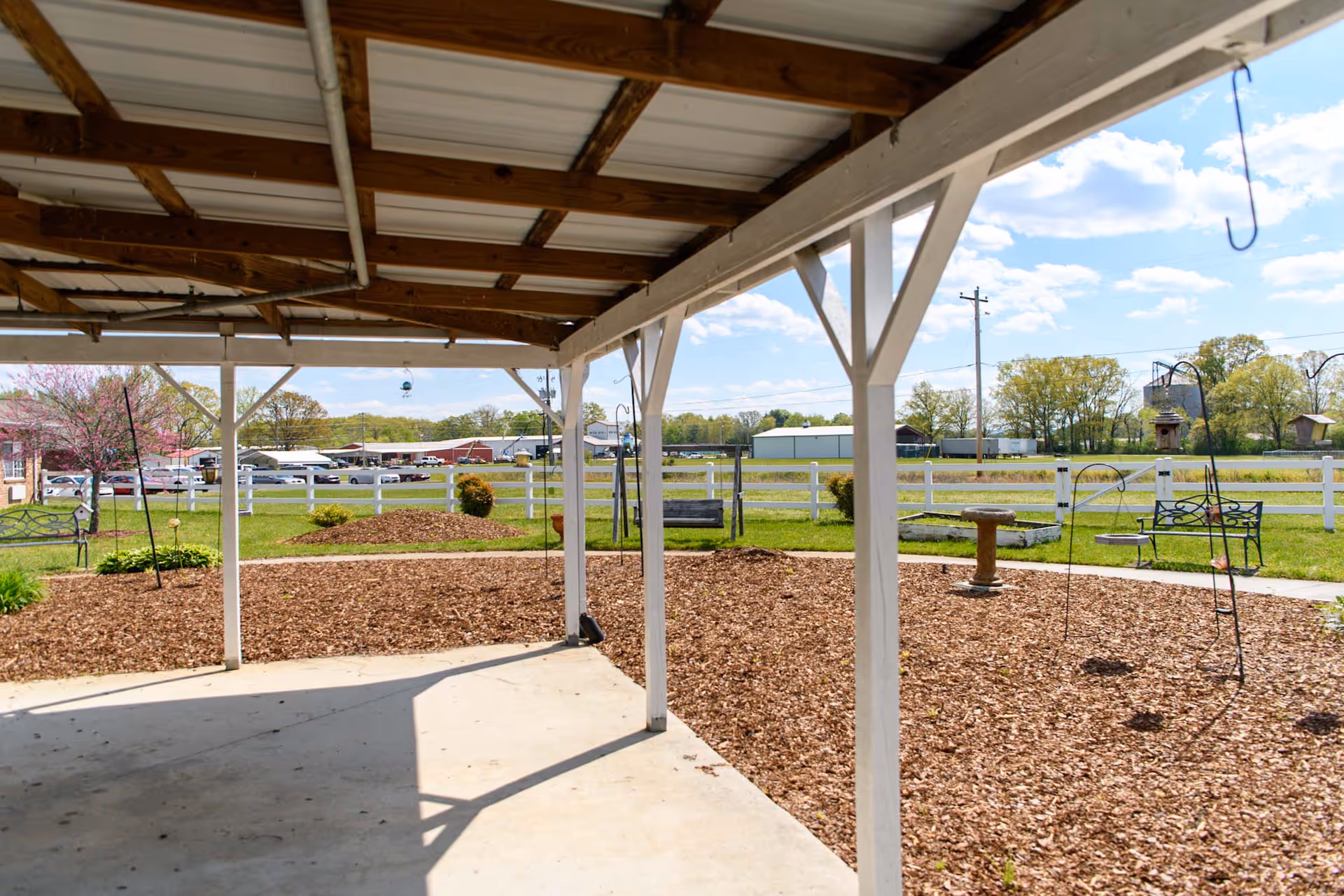 View from under a covered patio looking out onto a garden area with wood chip ground cover, bird feeders, benches, and a white fence. Beyond the fence, there are trees, buildings, and a partly cloudy sky.
