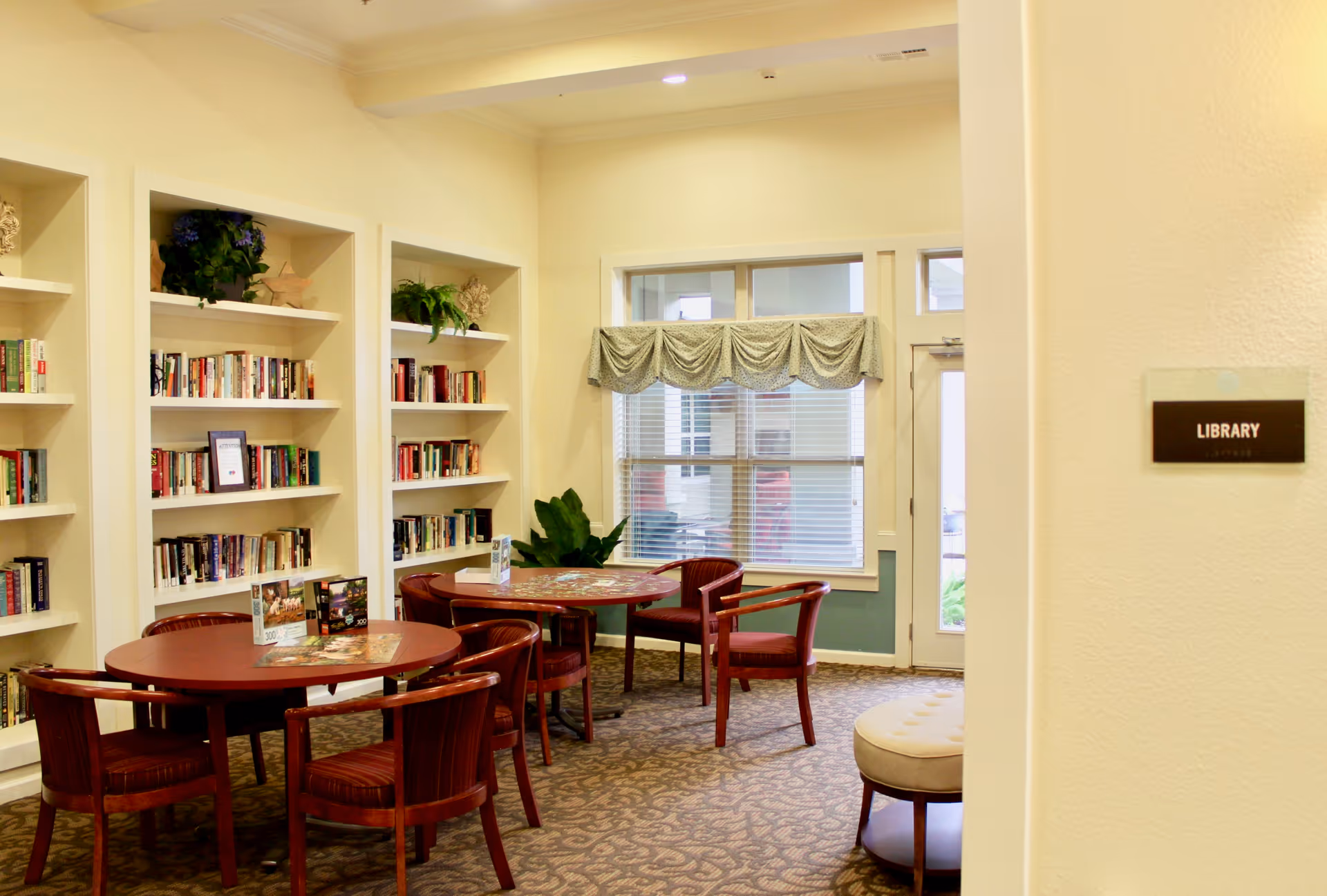 A cozy library room with built-in bookshelves filled with books and decorative plants. There are two round wooden tables surrounded by wooden chairs with red cushions. A window with blinds and a decorative valance lets in natural light. A small cushioned stool is visible near the entrance, and a sign on the wall reads 'LIBRARY'.