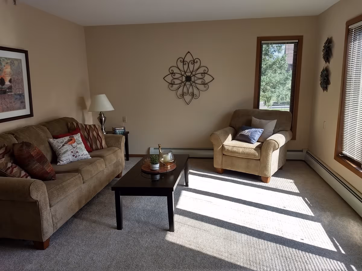 A cozy living room with a beige sofa and armchair, both adorned with decorative pillows. A black coffee table sits in the center with small decorative items on it. The room has beige walls with a framed picture and a metal wall decoration. Two windows with blinds allow natural light to stream in, casting shadows on the carpeted floor.