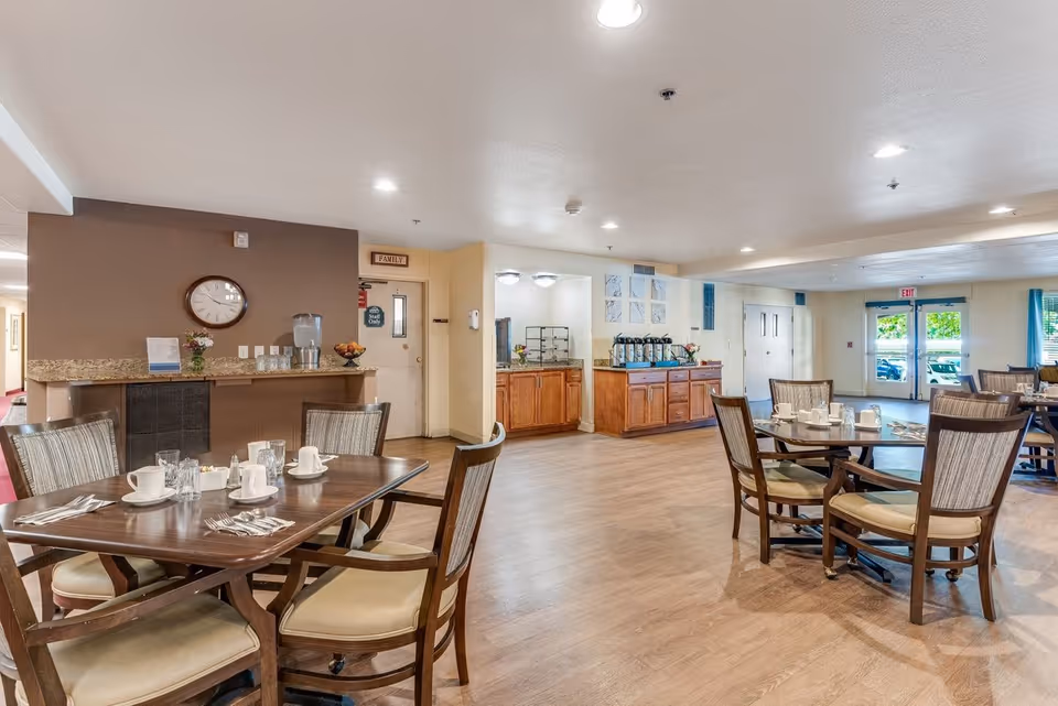 A spacious dining area in a senior living facility with wooden tables and cushioned chairs arranged neatly. Tables are set with cups, glasses, and utensils. The room features wood flooring, a beverage station with coffee urns and water dispenser, and large windows letting in natural light.