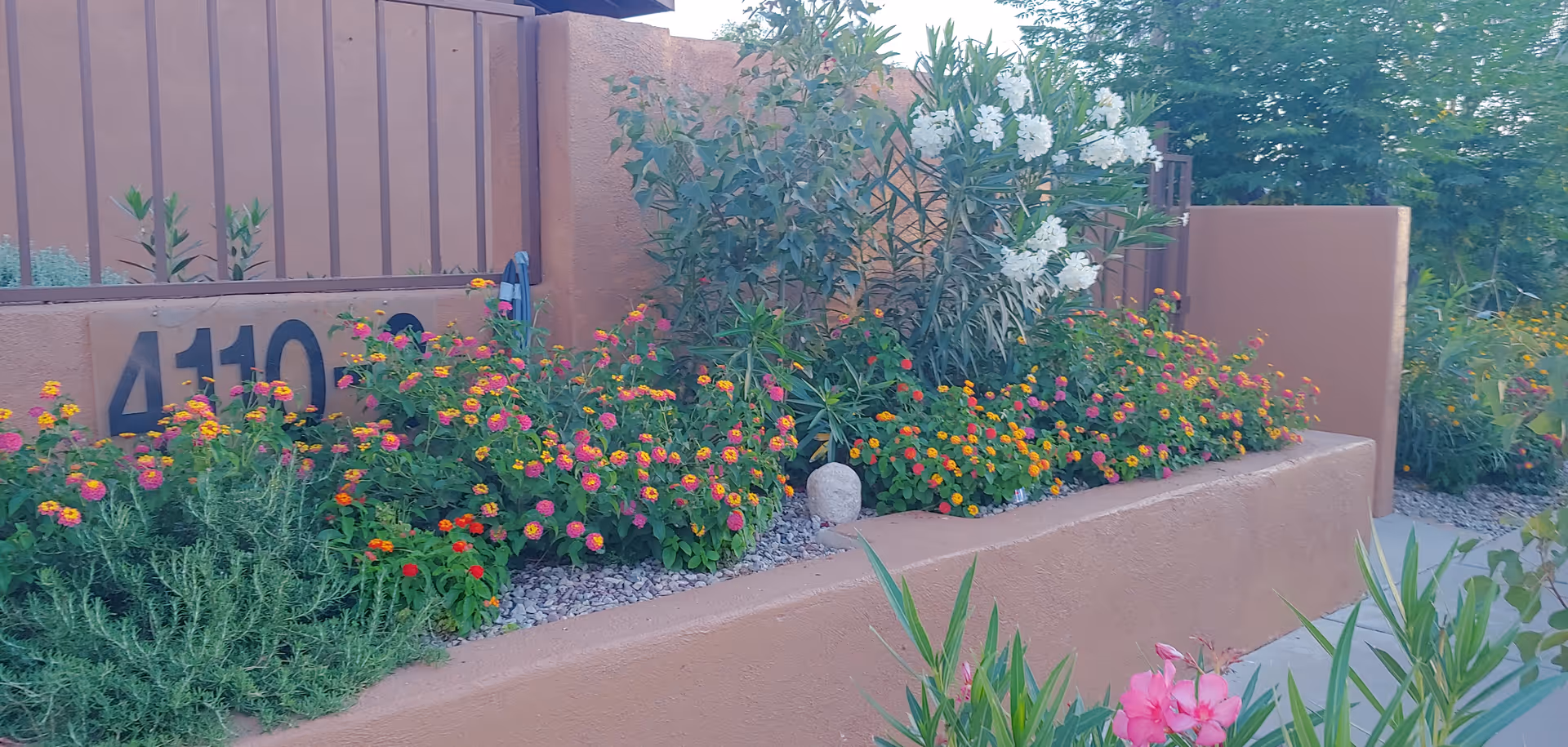 A landscaped outdoor garden area with colorful flowers and green shrubs planted in a raised bed made of brown stucco walls. The number 4110 is visible on the wall behind the flowers.