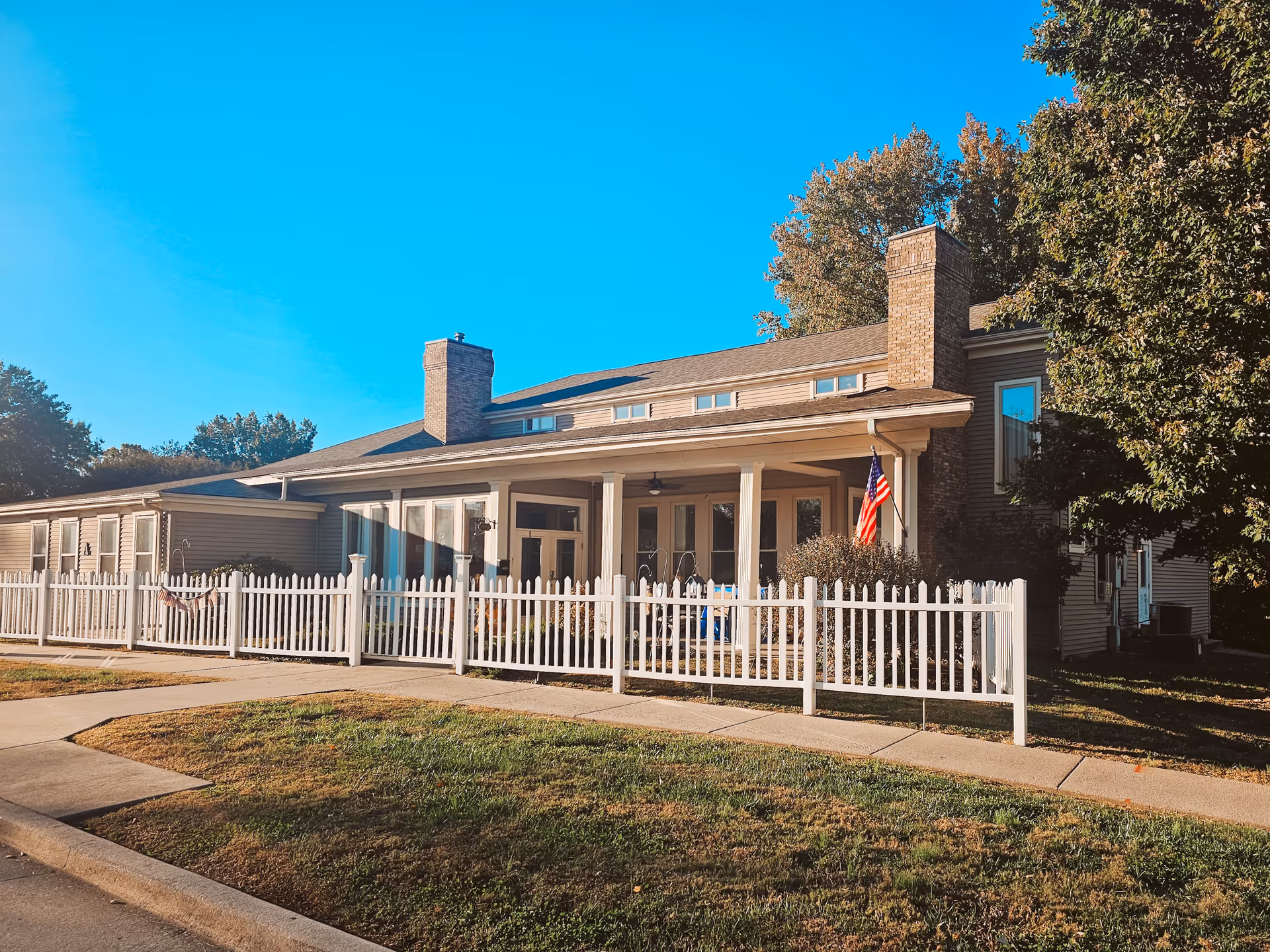 Exterior view of a single-story assisted living facility building with beige siding, two brick chimneys, a white picket fence, and an American flag near the entrance. The building is surrounded by trees and grass under a clear blue sky.
