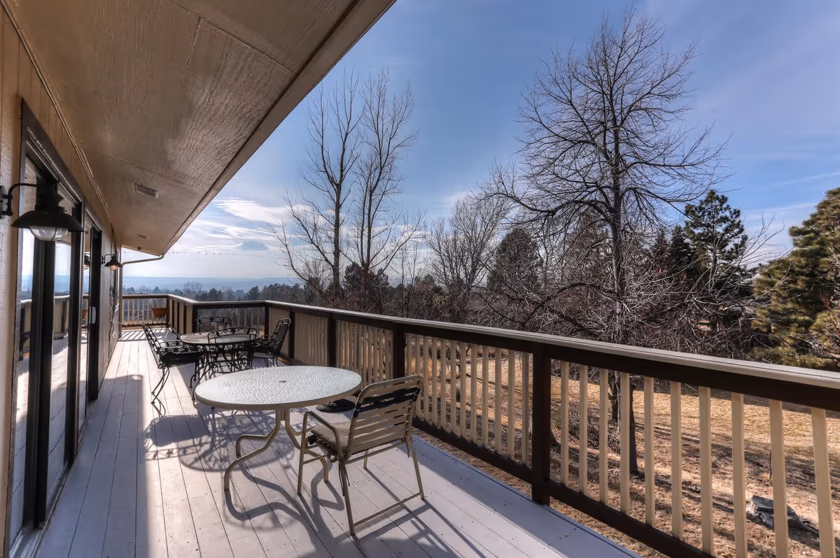Outdoor balcony area with several tables and chairs, overlooking a landscape with leafless trees and a clear blue sky.