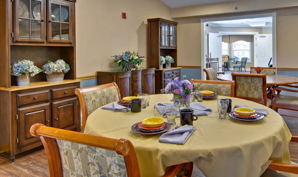 A dining room in a senior living facility with a round table covered with a yellow tablecloth, set with plates, bowls, cups, glasses, and napkins. There is a floral centerpiece on the table. The room has wooden furniture including cabinets with decorative plants and another table with chairs in the background. The space is well-lit with natural light coming from windows in an adjoining room.