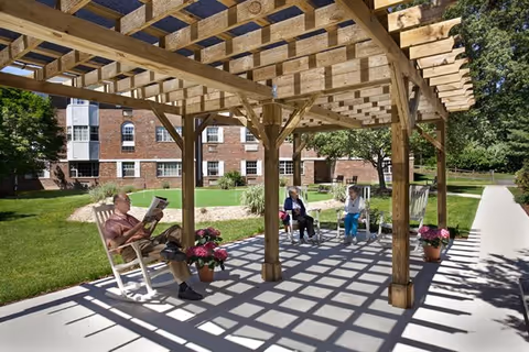 Three elderly people sitting under a wooden pergola in a garden area of a retirement community. One person is reading a newspaper while the other two are seated on a bench and a chair, engaged in conversation. The background shows a brick building and green lawn with trees and potted flowers around the seating area.