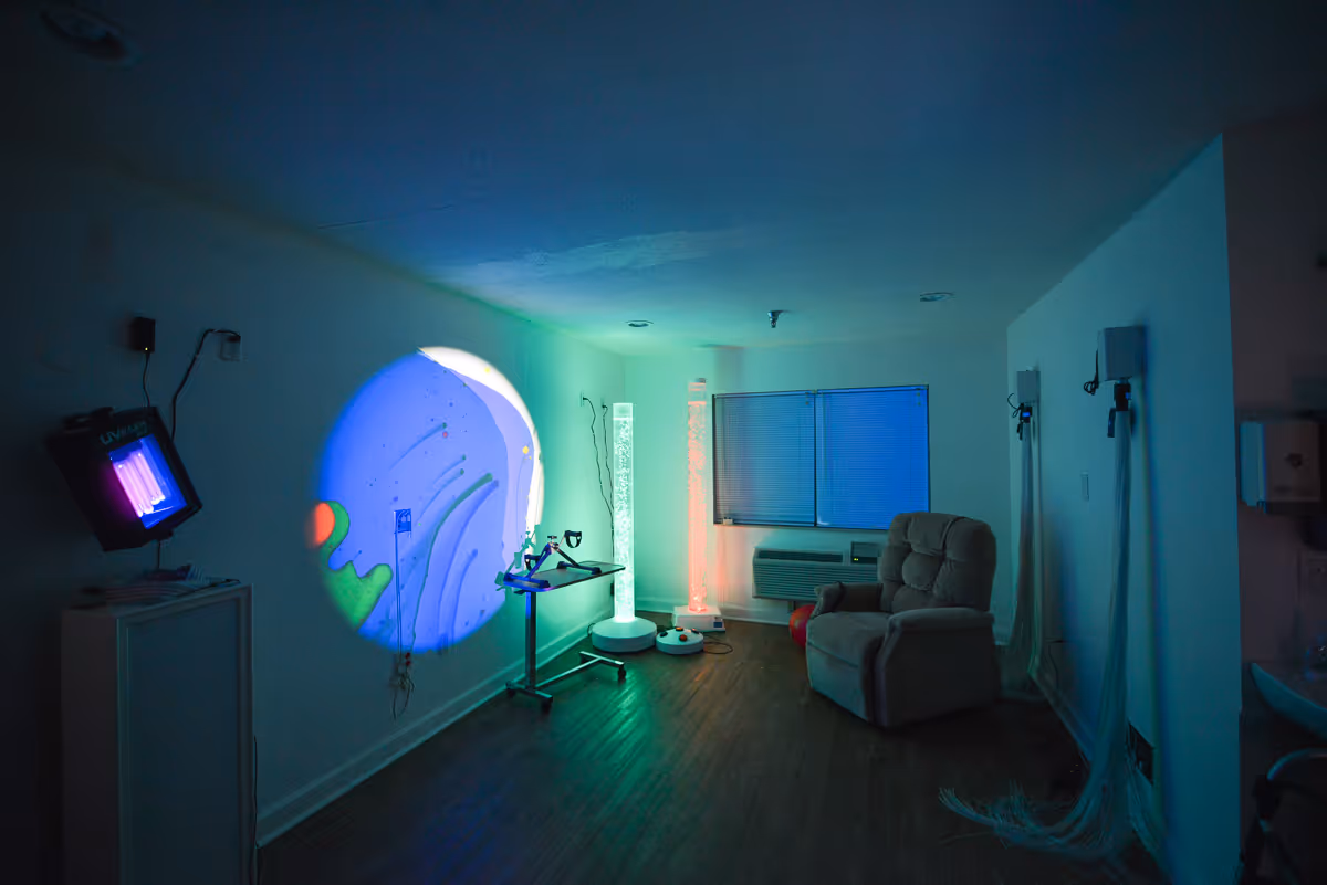 Dimly lit interior sensory/therapy room with colorful light projections on the wall, a recliner, and various therapeutic equipment.