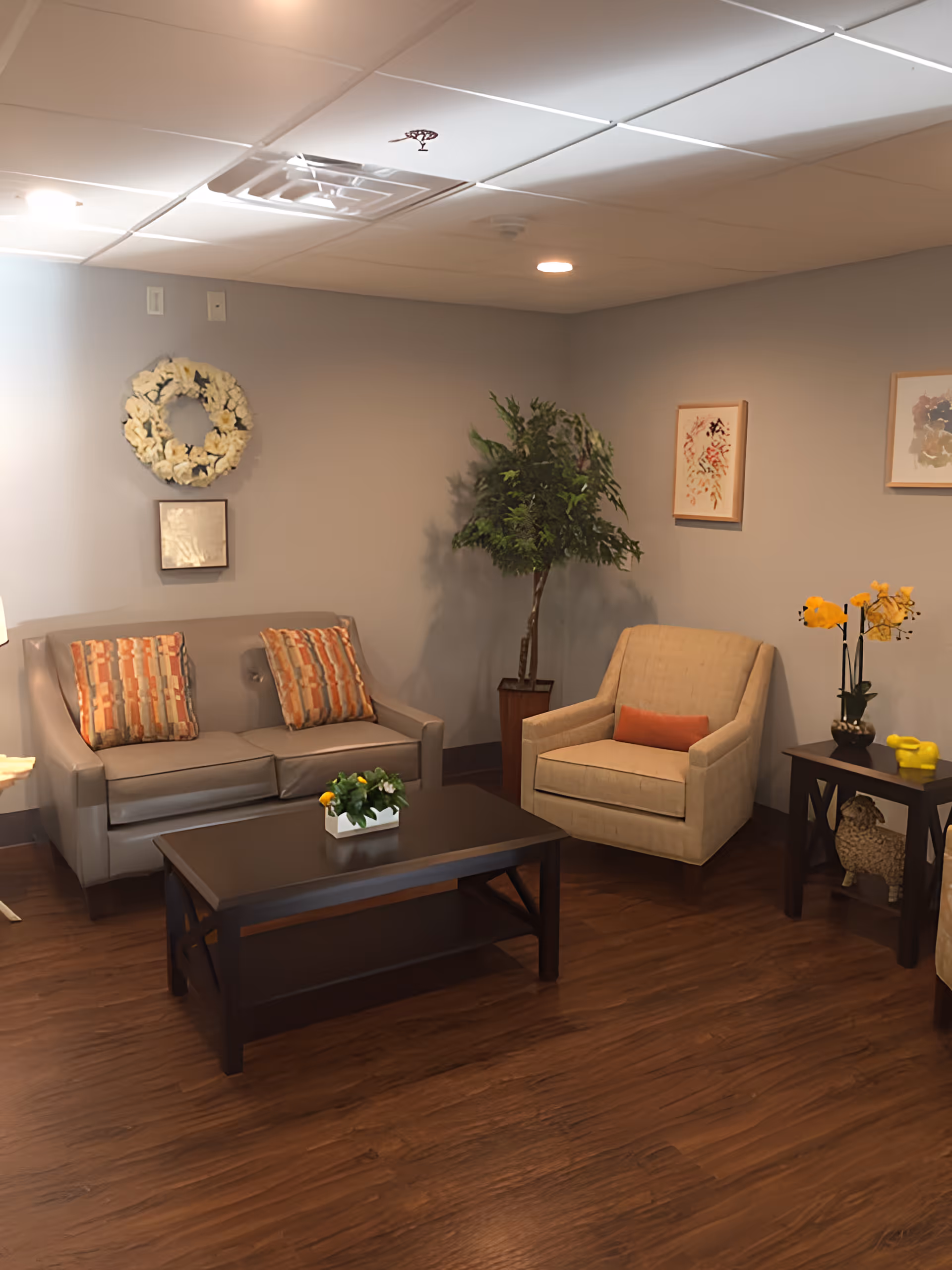 A cozy seating area with a sofa, armchair, coffee table, potted plant, and wall decorations in a senior living common room.