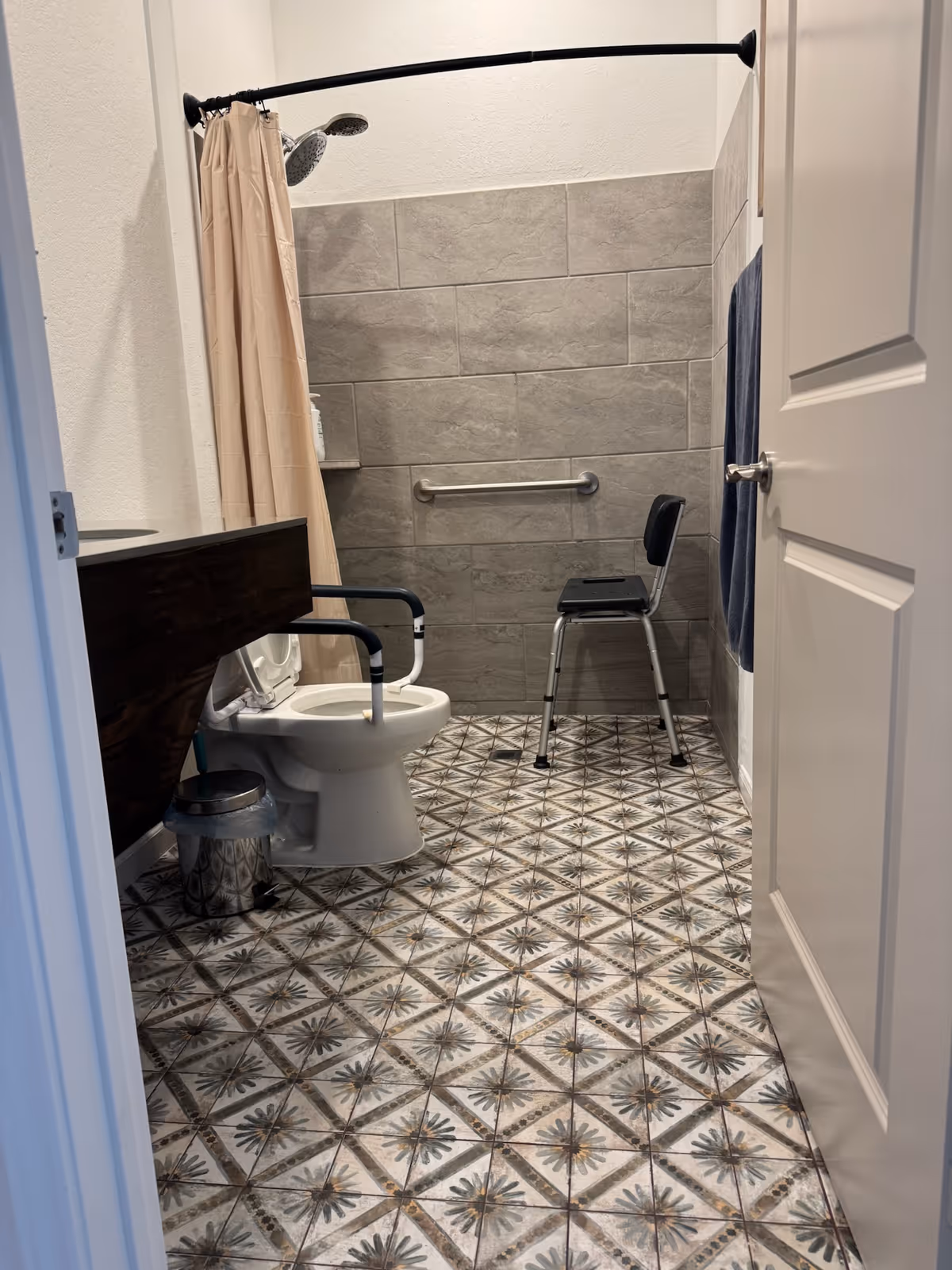 A bathroom with patterned tile floor, a white toilet with black safety rails, a metal trash can, a shower area with a beige curtain and dual shower heads, a black shower chair, and a towel hanging on the wall near the door.