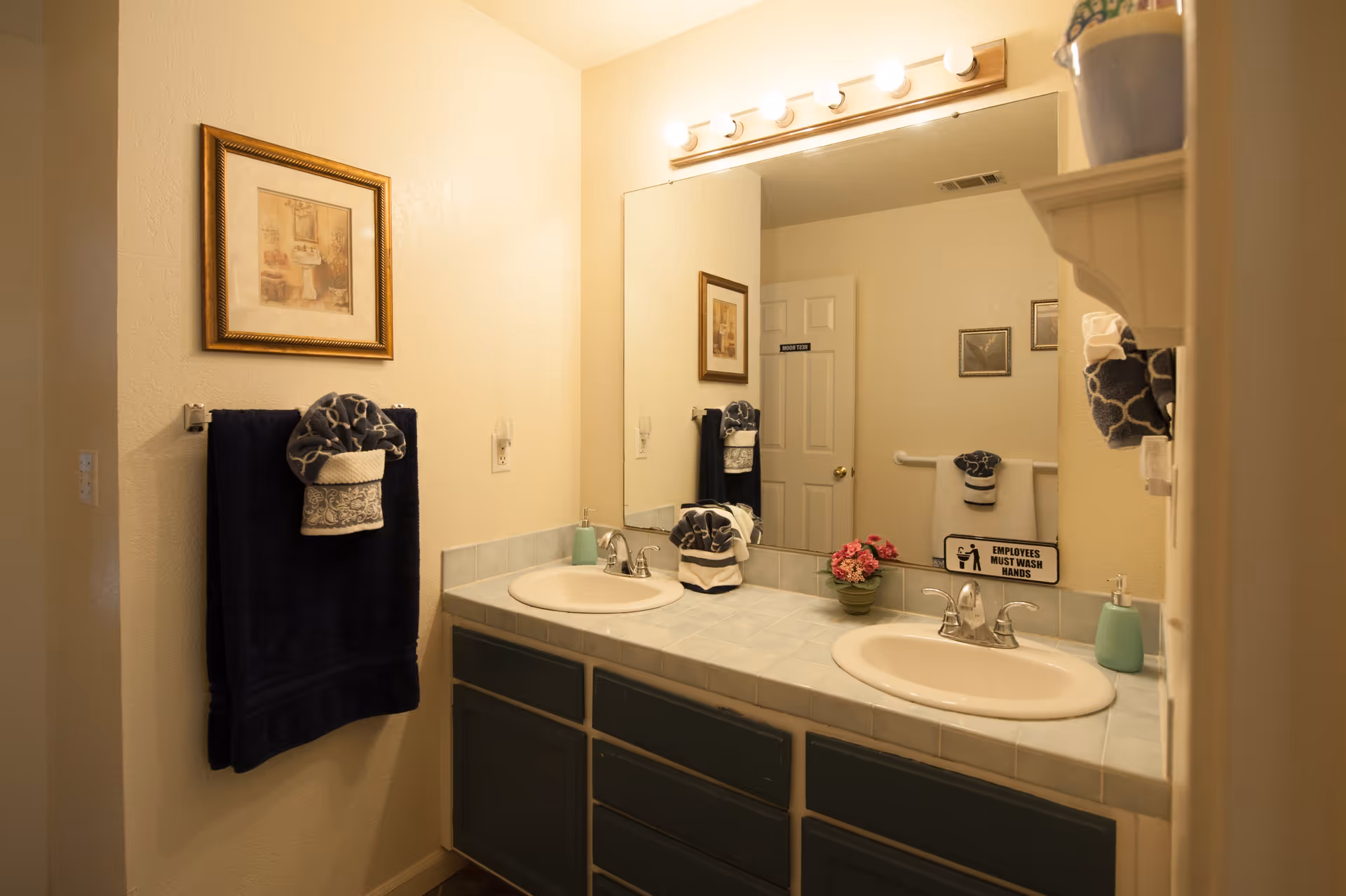 A bathroom with a double sink vanity featuring light blue tiled countertops and dark blue cabinets. Above the sinks is a large mirror with a row of lights. On the left wall, there is a framed picture and a towel rack holding a dark blue towel with a decorative folded towel on top. On the countertop, there is a small potted plant with pink flowers and a soap dispenser. In the mirror reflection, a door and additional framed pictures are visible, along with a sign that reads 'EMPLOYEES MUST WASH HANDS'.