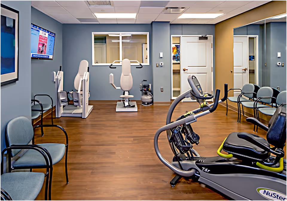A fitness room with exercise equipment including a recumbent elliptical machine and two seated strength training machines. The room has wood flooring, blue-gray walls, several chairs along the walls, a wall-mounted TV, and a large mirror on one side.