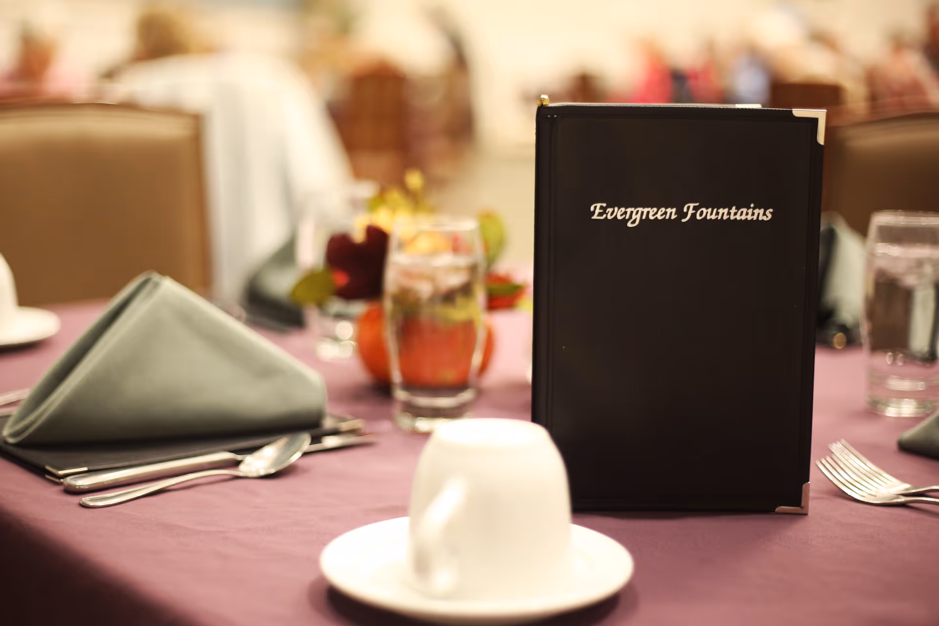 A close-up view of a dining table set with a purple tablecloth, a folded green napkin, silverware, a white cup and saucer, a glass of water, and a black menu book with the text 'Evergreen Fountains' on the cover. The background shows blurred chairs and other tables.