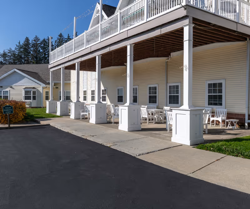 Outdoor covered patio area with white wooden chairs and tables arranged under a balcony supported by white pillars, adjacent to a beige building with multiple windows. A small green sign reads 'Tour Parking Only'.