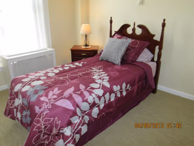 Single bed with a purple floral comforter and wooden headboard beside a nightstand and lamp in a small bedroom.