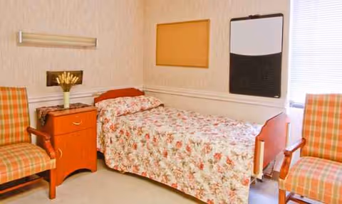 A bedroom in a senior care facility with floral bedspread, two plaid chairs, and a dresser.