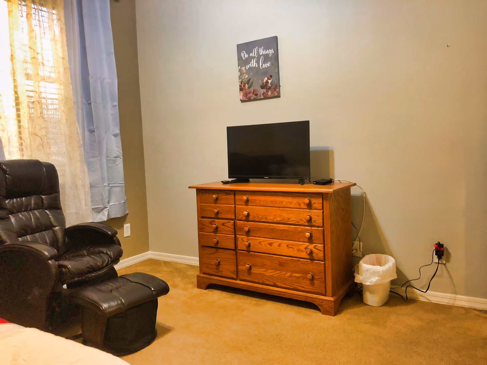 A cozy room with a wooden dresser holding a flat-screen TV, a black leather recliner chair with a matching footrest, beige carpet, and a window with sheer and blue curtains. A small trash can is placed next to the dresser, and a decorative wall art with the phrase 'Do all things with love' hangs above the TV.