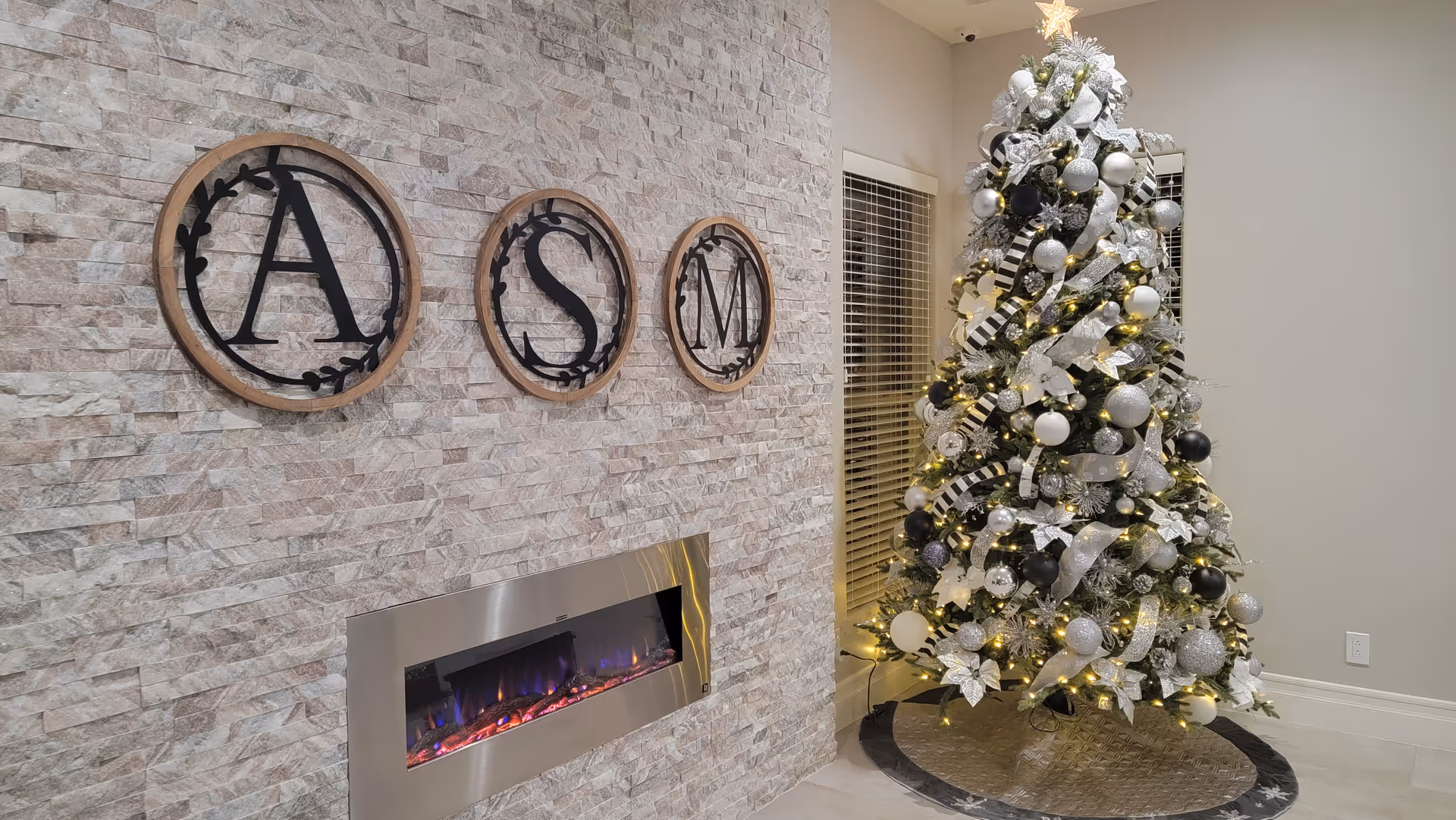 Decorated Christmas tree beside a stone accent wall with circular "A S M" letters and a modern electric fireplace.