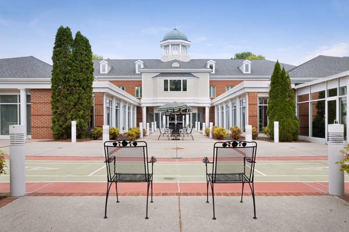 Outdoor courtyard area of The Colonnades facility featuring two black metal chairs in the foreground, a shuffleboard court, and a central patio with tables and umbrellas. The building has large windows, brick and siding exterior, and a cupola on the roof under a clear blue sky.