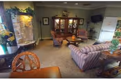 A cozy living room area with striped sofas, armchairs, a wooden cabinet with glass doors, a small TV mounted on the wall, and various decorative plants and flowers. The room has carpeted flooring and soft lighting, creating a warm and inviting atmosphere.