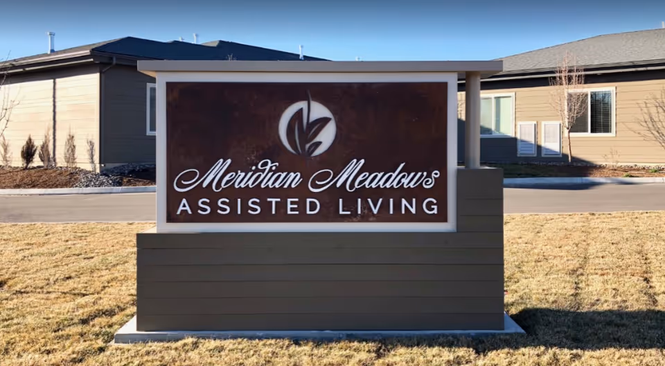 Outdoor view of a sign for Meridian Meadows Assisted Living placed on a grassy area with buildings in the background under clear daylight.