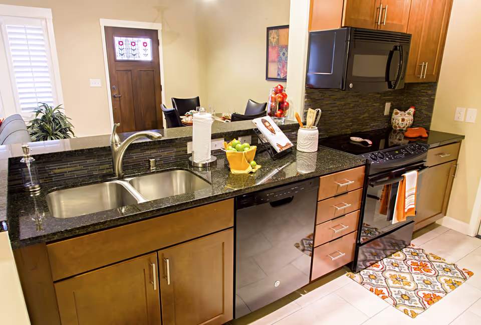 Modern kitchen with dark granite countertops, a double stainless steel sink, a black dishwasher, and a black stove with a microwave above it. The kitchen has wooden cabinets and drawers, a colorful patterned rug on the floor, and various kitchen items like a paper towel holder, a bowl of green apples, and utensils in a container. In the background, there is a dining area with a table and chairs, a wooden front door with decorative glass panels, and a plant near a window with white shutters.