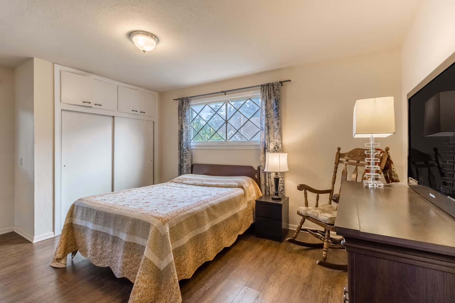 A cozy bedroom with a bed covered in a beige and floral quilt, a wooden rocking chair, a dark wooden dresser with a lamp, a nightstand with a lamp, and a window with patterned curtains letting in natural light.