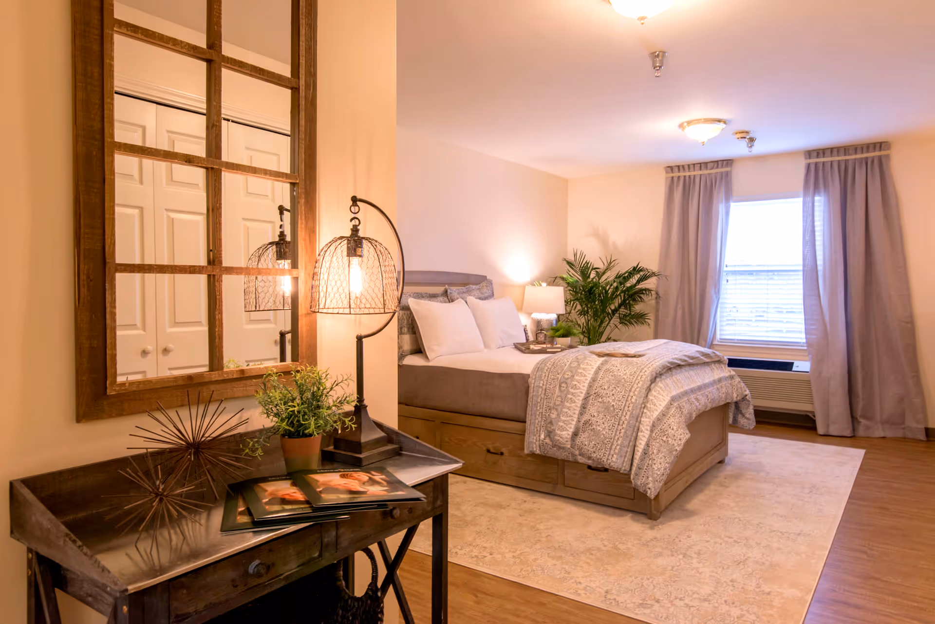 A cozy bedroom with a neatly made bed featuring white pillows and a patterned comforter. There is a wooden nightstand with a lamp and a green plant next to the bed. A large window with curtains allows natural light to enter the room. In the foreground, there is a wooden desk with a decorative lamp, a small potted plant, and some magazines. A large wooden-framed mirror hangs on the wall above the desk. The room has warm lighting and a light-colored rug on the wooden floor.