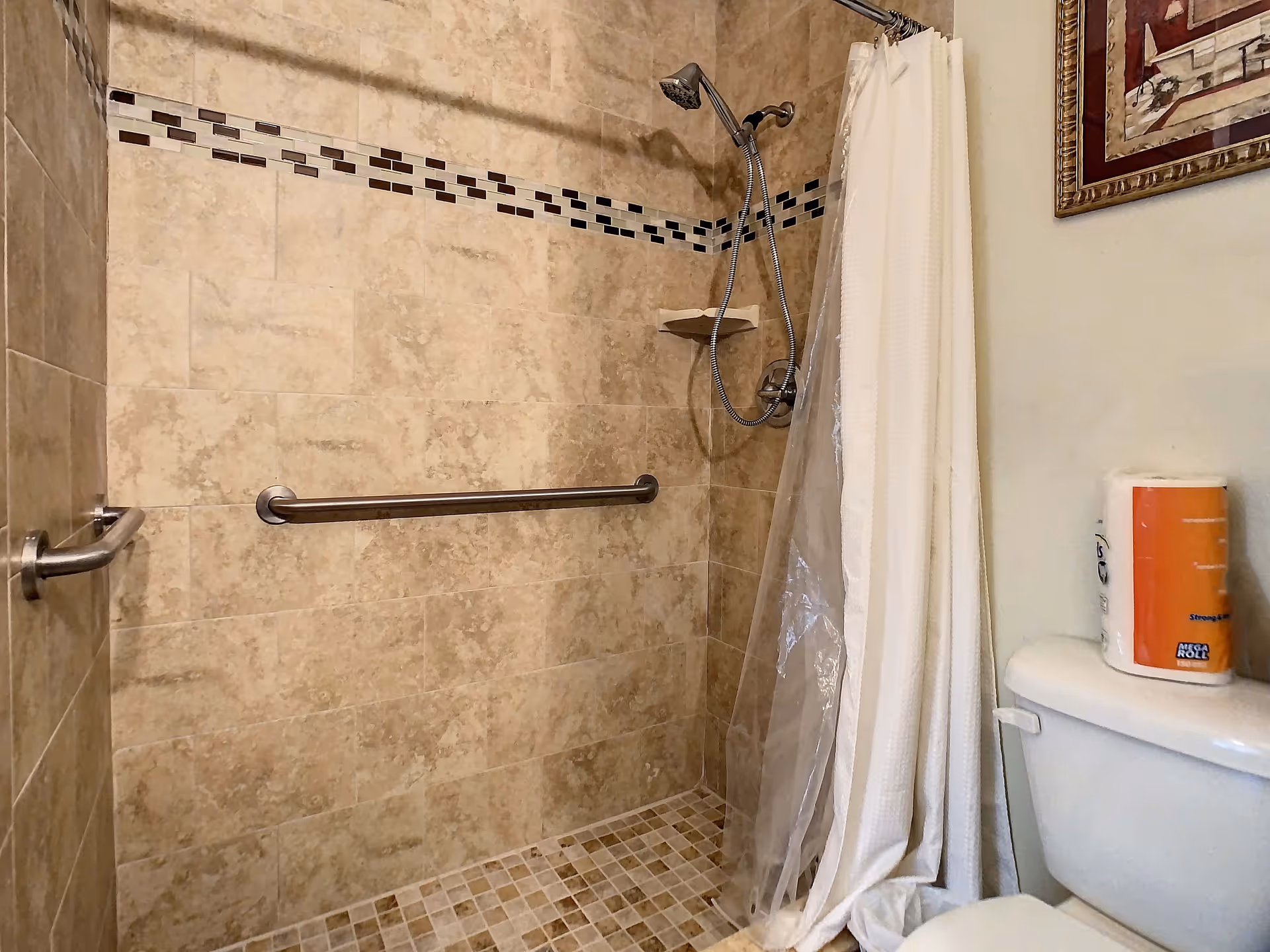 A tiled shower area with beige and brown mosaic tiles on the floor and a decorative horizontal strip of small tiles on the wall. The shower has a handheld showerhead attached to a flexible hose and a metal grab bar on the wall. A white shower curtain is partially drawn to the side, and a toilet with a roll of paper towels on top is visible next to the shower. A framed picture hangs on the wall above the toilet.