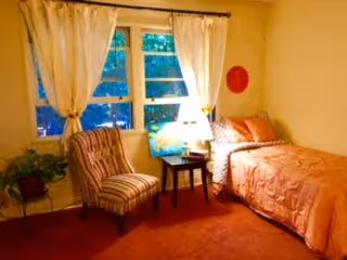 Small, warmly lit bedroom with a single bed, bedside table and lamp, a striped armchair, potted plant, and a curtained window.