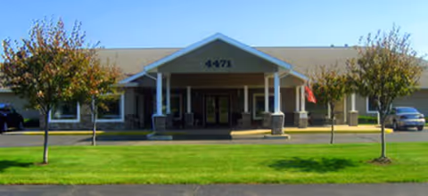 Front exterior view of a single-story building with a covered entrance supported by columns, a green lawn, small trees, and a few parked cars. The building has the number 4471 displayed above the entrance.