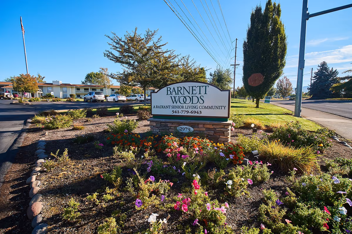 Entrance sign for Barnett Woods senior living community surrounded by flowerbeds with the facility and parked cars in the background.