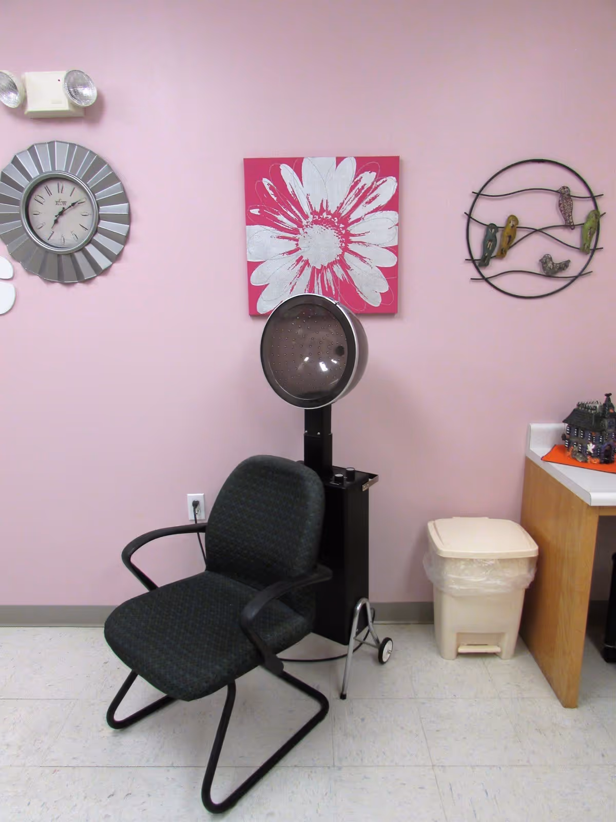A small room with pink walls featuring a black salon chair in front of a vintage hair dryer. On the wall above the dryer, there is a large clock, a pink and white flower painting, and a metal wall decoration with birds. A trash can is placed next to a wooden counter with a small decorative house on it.