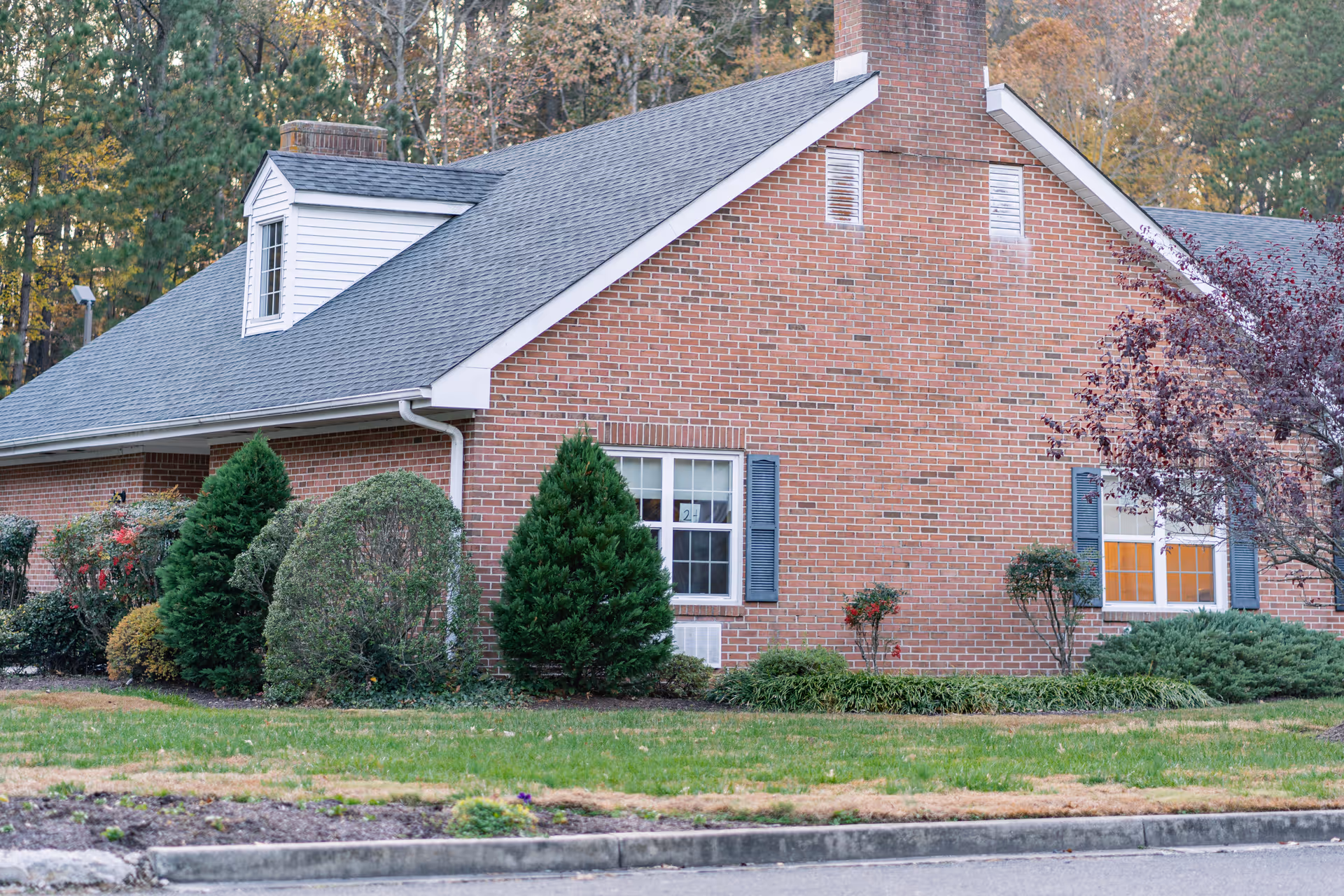Exterior view of a single-story brick building with a gray shingled roof, white window frames, and blue shutters. The building is surrounded by neatly trimmed bushes and trees, with a grassy lawn in front and a paved driveway or road in the foreground. Trees with autumn foliage are visible in the background.