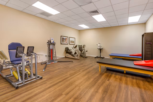 A fitness and rehabilitation room with exercise equipment including stationary bikes, a leg press machine, and therapy tables. The room has wood flooring, beige walls, and a white drop ceiling with fluorescent lighting. Two framed pictures hang on the wall.