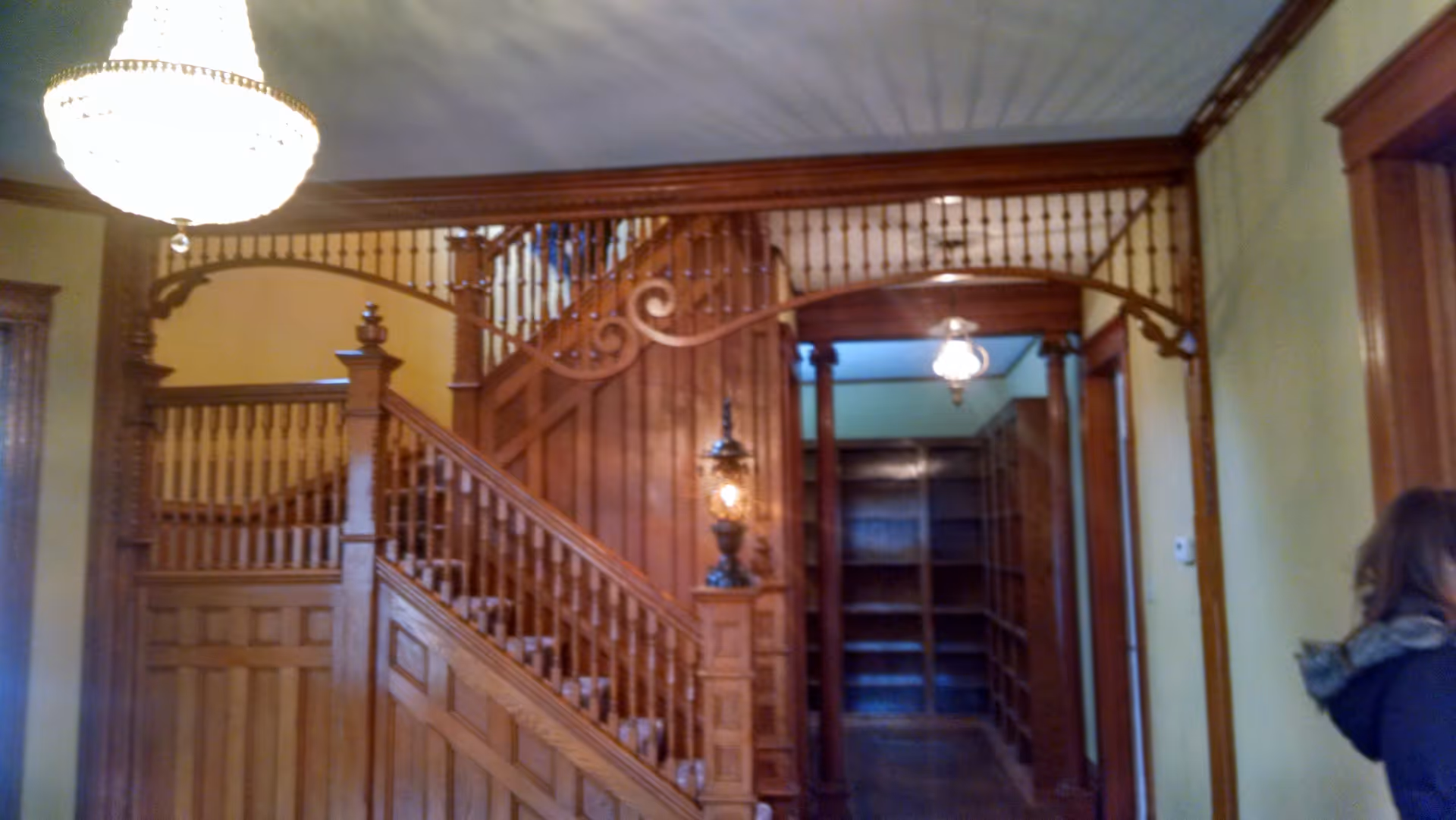 Interior view of a senior living facility showing a wooden staircase with ornate railing and wood paneling. There is a hanging chandelier light fixture above and a hallway with built-in wooden shelves in the background. A person wearing a dark jacket is partially visible on the right side of the image.