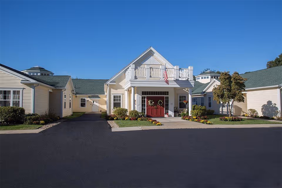 Front entrance of a single-story yellow senior living building with a covered porch, red double doors, an American flag, and landscaped flower beds under a clear blue sky.