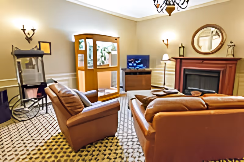 A cozy living room with brown leather sofas arranged around a wooden coffee table. There is a fireplace with a round mirror above it on the right wall, a flat-screen TV on a wooden stand, and a wooden cabinet with glass doors. The room is softly lit with wall sconces and a chandelier, and the floor is covered with a patterned carpet.