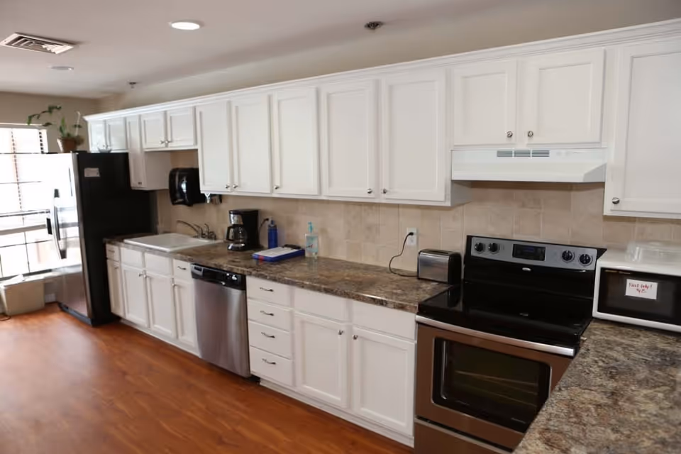 A clean and modern kitchen with white cabinets, a stainless steel refrigerator, dishwasher, stove, and a microwave. The countertop is a dark marble pattern, and the floor is wooden. There is a coffee maker, toaster, and hand sanitizer on the counter, with a window letting in natural light.