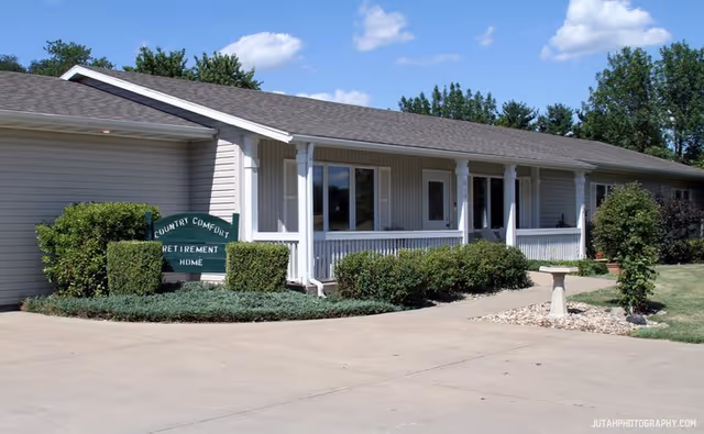Exterior view of Country Comfort Retirement Home, a single-story building with a covered porch, white columns, and surrounded by bushes and greenery under a blue sky with some clouds.