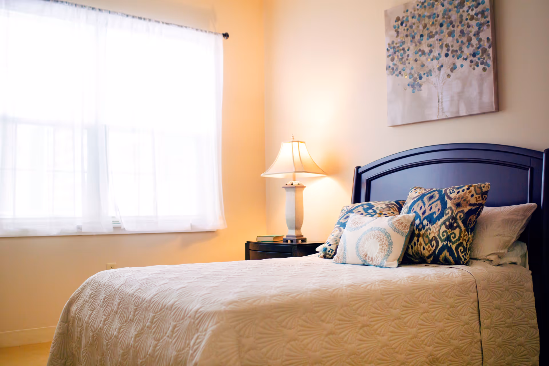 A cozy bedroom with a neatly made bed featuring a white quilted bedspread and several decorative pillows in blue and beige patterns. A dark wooden headboard is visible, along with a bedside table holding a lamp and a couple of books. A window with sheer white curtains allows natural light to brighten the room. A painting of a tree with blue and beige leaves hangs on the wall above the bed.
