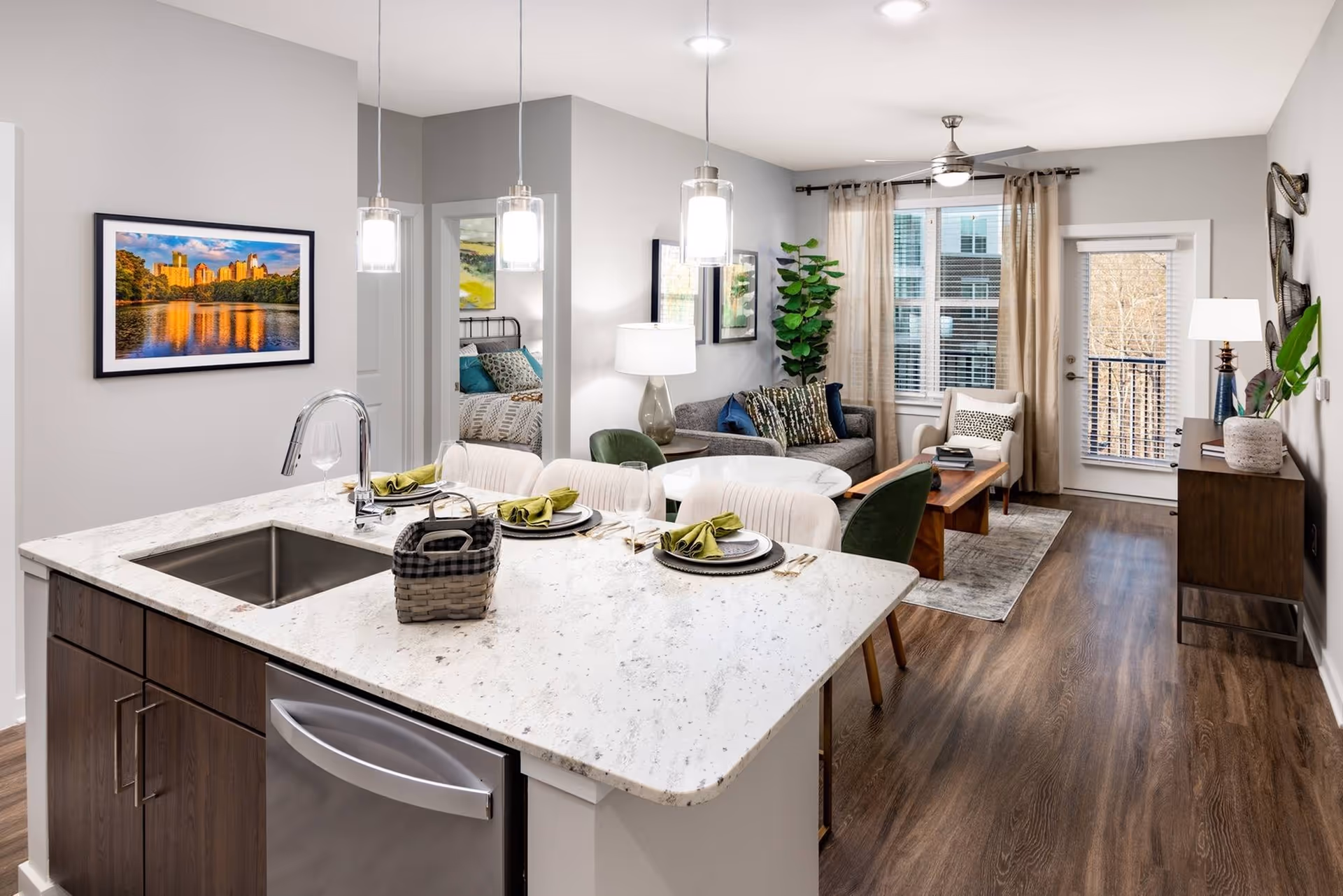 Modern open-concept living space with a kitchen island featuring a sink and place settings for four, a dining table with chairs, a cozy living room with a sofa, armchair, coffee table, and large window with curtains. A bedroom is visible through an open door. The room has wooden flooring and neutral-colored walls with framed artwork and decorative plants.