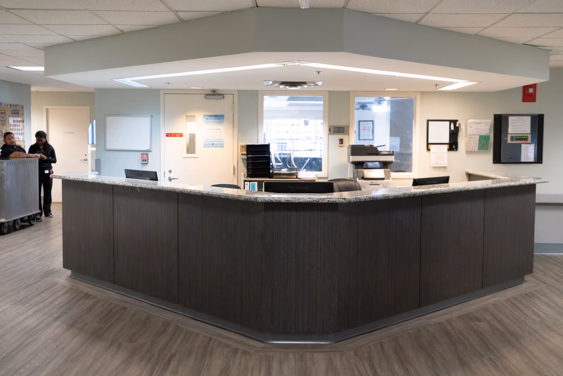 Reception desk area inside a healthcare facility with a dark wood counter and granite top. Behind the desk are office equipment, windows, and doors. Two staff members are seen on the left side near a cart.