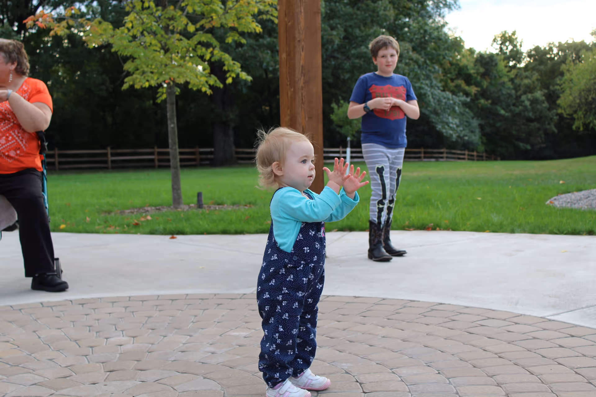 A toddler wearing a blue long-sleeve shirt and patterned overalls stands on a circular paved area outdoors, clapping hands. In the background, a young boy in a blue shirt and striped pants with a skeleton design stands on a concrete path, and a woman in an orange shirt sits on a bench near a tree. The scene is set in a green park-like area with grass, trees, and a wooden fence.